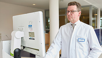 Ein Arzt im weißen Kittel steht an einer mobilen Monitorstation und betrachtet medizinische Bilddaten im Klinikflur.