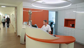 Pflegekraft sitzt hinter einem geschwungenen Empfangstresen, im Hintergrund ein heller Flur mit weiteren Räumen.