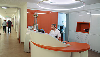 Pflegekraft sitzt hinter einem geschwungenen Empfangstresen, im Hintergrund ein heller Flur mit weiteren Räumen.