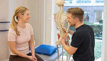 Ein Physiotherapeut zeigt an einem Skelettmodell die Wirbelsäule, während eine Patientin auf einer Behandlungsliege sitzt und aufmerksam zuhört.