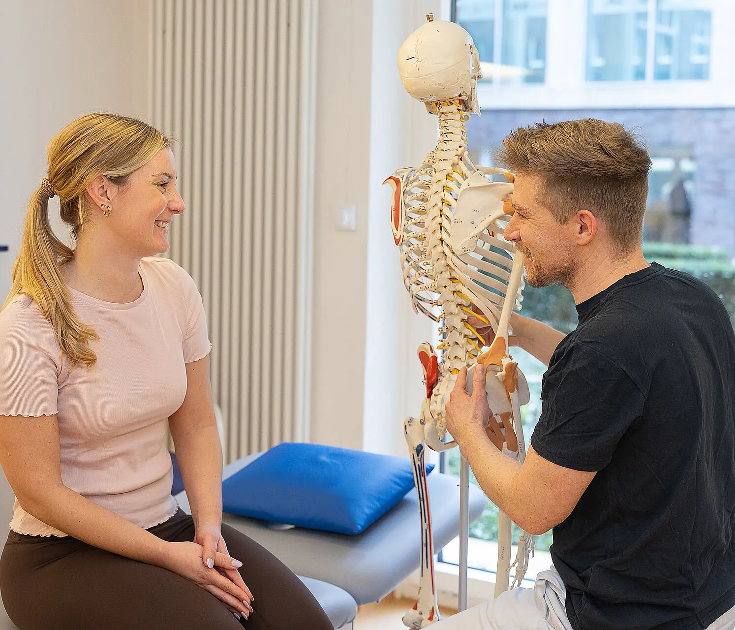 Ein Physiotherapeut erläutert einer Patientin an einem Skelettmodell, wo die Probleme bzw. Schmerzen in der Wirbelsäule ihren Ursprung haben.
