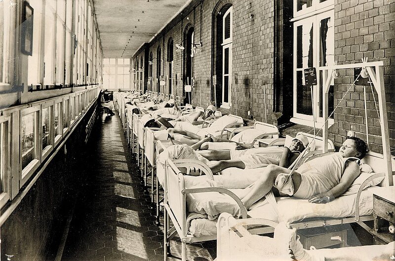 Historische Aufnahme einer Liegehalle mit mehreren Patient:innen in Betten entlang eines langen Flurs mit großen Fenstern.
