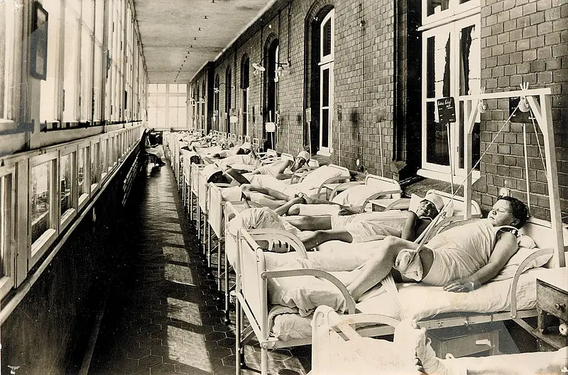 Historische Aufnahme einer Liegehalle mit mehreren Patient:innen in Betten entlang eines langen Flurs mit großen Fenstern.