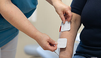 Eine Pflegefachperson bringt zwei Elektroden eines TENS-Geräts am Unterarm einer sitzenden Person an, die über Kabel mit dem Gerät verbunden sind.