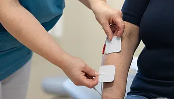 Eine Pflegefachperson bringt zwei Elektroden eines TENS-Geräts am Unterarm einer sitzenden Person an, die über Kabel mit dem Gerät verbunden sind.