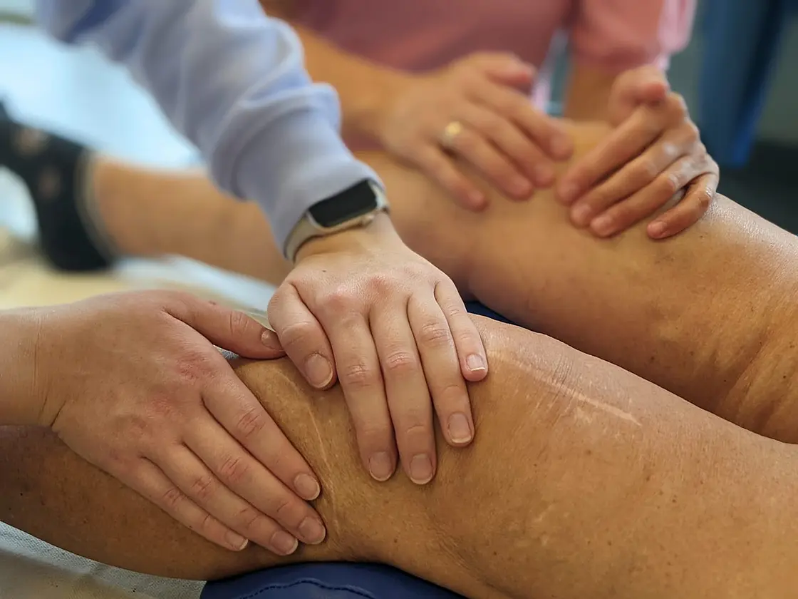 Mehrere Hände führen an einem ausgestreckten Bein manuelle Techniken entlang einer längeren Operationsnarbe durch. Die Behandlung findet auf einer Therapieliege statt.