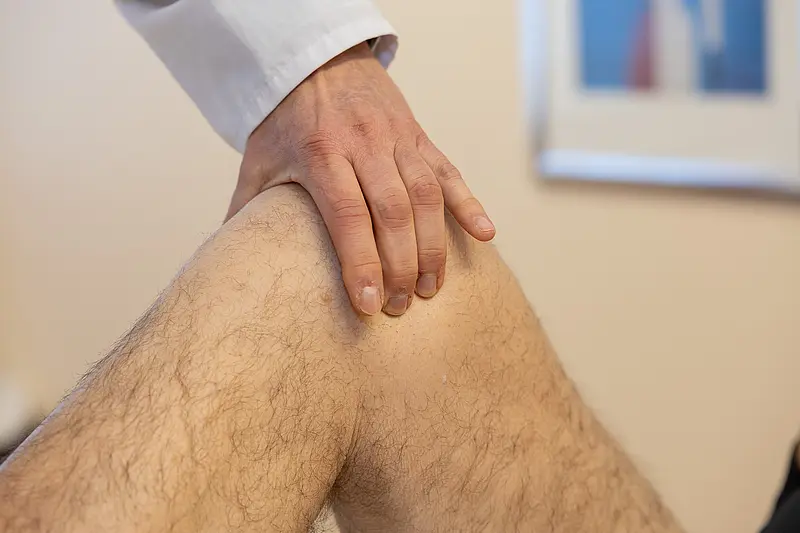 Eine Hand eines Arztes liegt auf dem Knie eines Patienten während einer Untersuchung. Der Fokus liegt auf der Berührung und dem Knie.