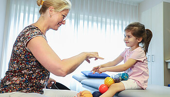 Eine Physiotherapeutin sitzt einem Kind gegenüber und leitet eine spielerische Übung auf der Behandlungsliege an; das Kind hält verschiedene Therapiebälle auf dem Schoß.