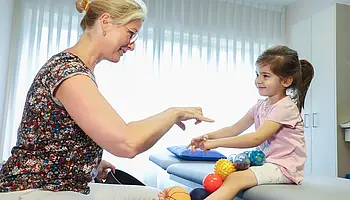 Eine Physiotherapeutin sitzt einem Kind gegenüber und leitet eine spielerische Übung auf der Behandlungsliege an; das Kind hält verschiedene Therapiebälle auf dem Schoß.