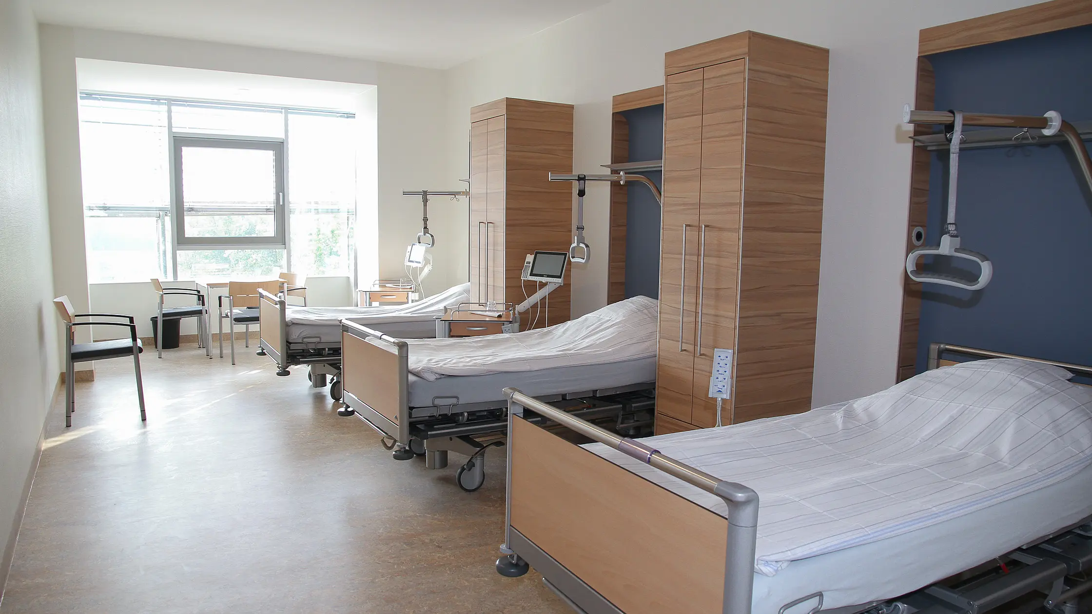 Mehrbettzimmer mit modernen Pflegebetten, Nachttischen und Sitzgelegenheiten, Tageslicht durch ein großes Fenster.