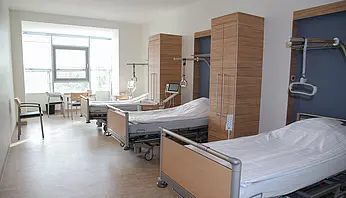 Mehrbettzimmer mit modernen Pflegebetten, Nachttischen und Sitzgelegenheiten, Tageslicht durch ein großes Fenster.