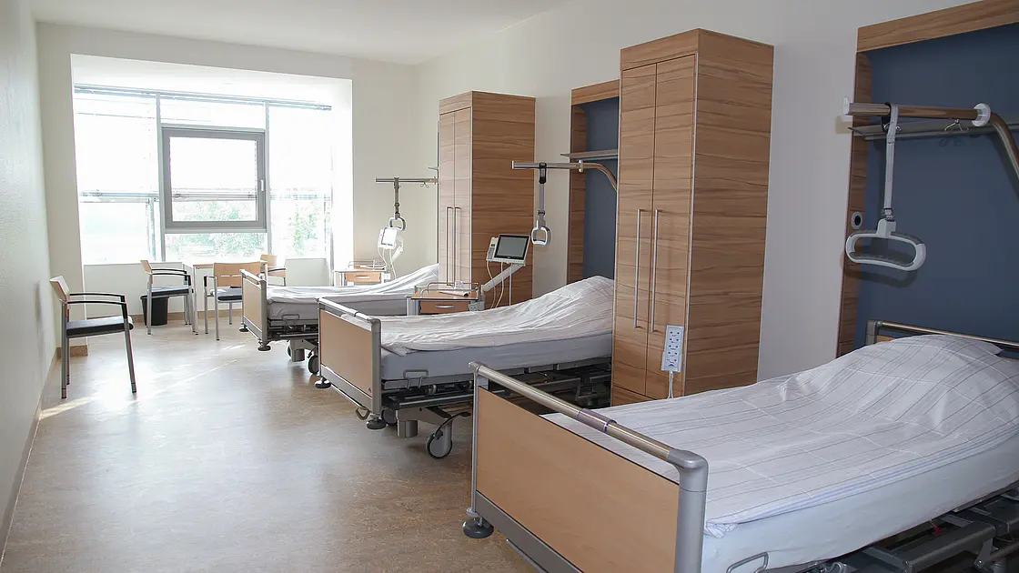 Mehrbettzimmer mit modernen Pflegebetten, Nachttischen und Sitzgelegenheiten, Tageslicht durch ein großes Fenster.