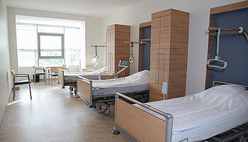 Mehrbettzimmer mit modernen Pflegebetten, Nachttischen und Sitzgelegenheiten, Tageslicht durch ein großes Fenster.