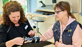 Eine Ergotherapeutin überprüft die Einstellung einer Handorthese am Arm einer Patientin, die den Arm auf einer gepolsterten Ablage abstützt.
