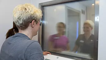 Eine Person steht vor dem Bedienpult der Kältekammer und blickt durch ein Fenster, hinter dem zwei weitere Personen zu sehen sind.