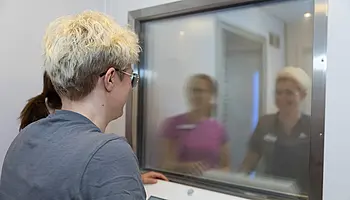 Eine Person steht vor dem Bedienpult der Kältekammer und blickt durch ein Fenster, hinter dem zwei weitere Personen zu sehen sind.