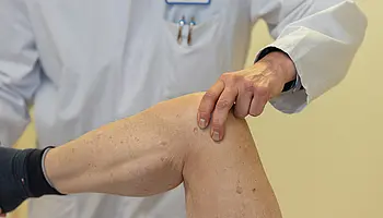 Eine Hand eines Arztes liegt auf dem Knie eines Patienten während einer Untersuchung. Der Fokus liegt auf der Berührung und dem Knie.