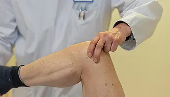 Eine Hand eines Arztes liegt auf dem Knie eines Patienten während einer Untersuchung. Der Fokus liegt auf der Berührung und dem Knie.