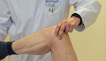 Eine Hand eines Arztes liegt auf dem Knie eines Patienten während einer Untersuchung. Der Fokus liegt auf der Berührung und dem Knie.