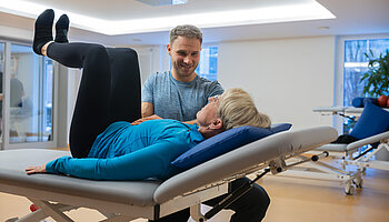 Eine Patientin liegt auf einer Therapieliege und führt eine Übung aus, während ein Therapeut sie anleitet.