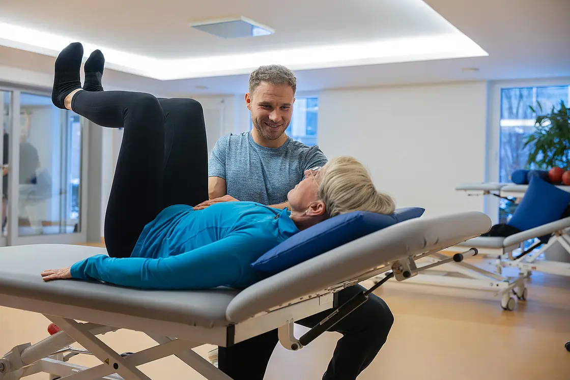 Eine Patientin liegt auf einer Therapieliege und führt eine Übung aus, während ein Therapeut sie anleitet.