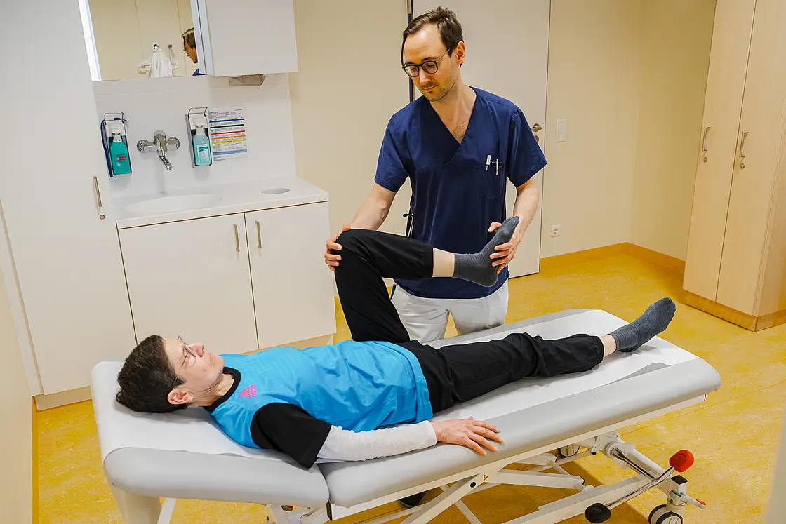 Behandelnde Person hebt das Bein einer auf der Untersuchungsliege liegenden Patientin zur Prüfung der Beweglichkeit an.