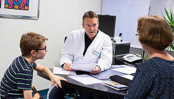 Arzt in weißem Kittel begrüßt einen Jungen in einem Behandlungsraum der Kinder- und Jugendrheumatologie.