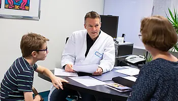 Arzt in weißem Kittel begrüßt einen Jungen in einem Behandlungsraum der Kinder- und Jugendrheumatologie.