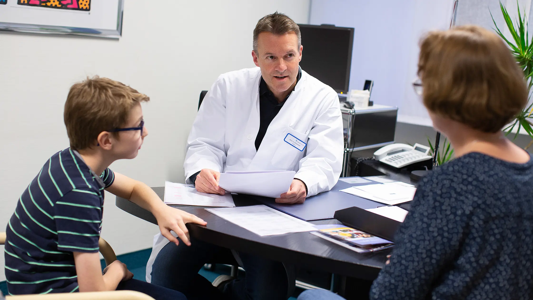 Arzt in weißem Kittel begrüßt einen Jungen in einem Behandlungsraum der Kinder- und Jugendrheumatologie.