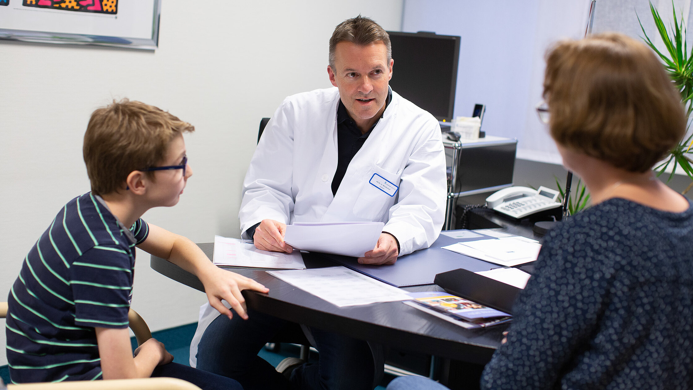 Arzt in weißem Kittel begrüßt einen Jungen in einem Behandlungsraum der Kinder- und Jugendrheumatologie.