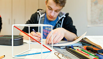 Jugendlicher sitzt an einem Tisch und arbeitet konzentriert an einer schriftlichen Aufgabe; Schulmaterialien liegen ausgebreitet vor ihm in einem Klassenraum der Klinikschule.