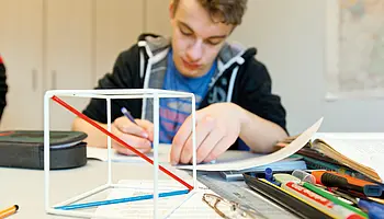 Jugendlicher sitzt an einem Tisch und arbeitet konzentriert an einer schriftlichen Aufgabe; Schulmaterialien liegen ausgebreitet vor ihm in einem Klassenraum der Klinikschule.