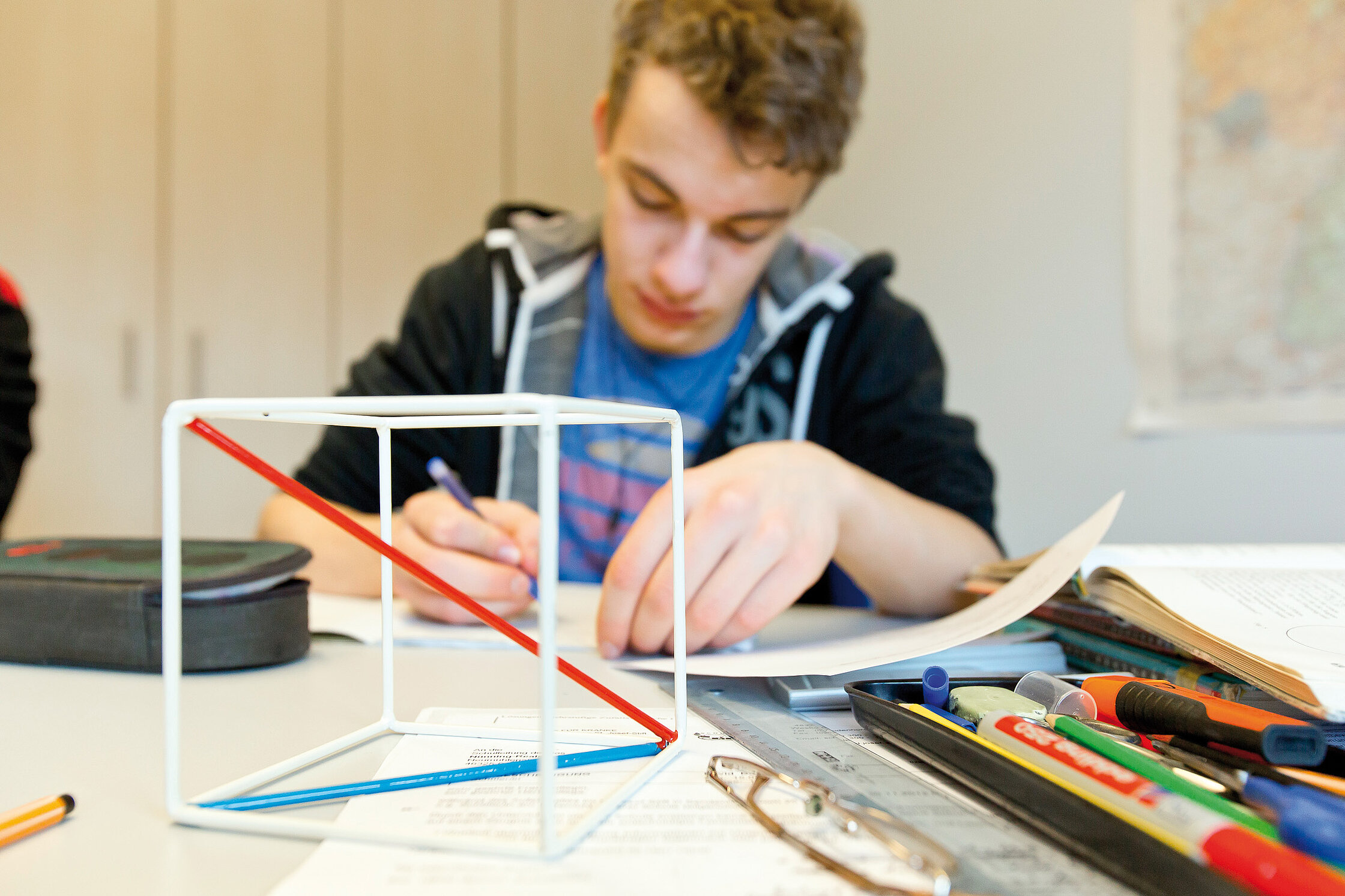 Jugendlicher sitzt an einem Tisch und arbeitet konzentriert an einer schriftlichen Aufgabe; Schulmaterialien liegen ausgebreitet vor ihm in einem Klassenraum der Klinikschule.
