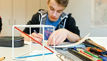 Jugendlicher sitzt an einem Tisch und arbeitet konzentriert an einer schriftlichen Aufgabe; Schulmaterialien liegen ausgebreitet vor ihm in einem Klassenraum der Klinikschule.