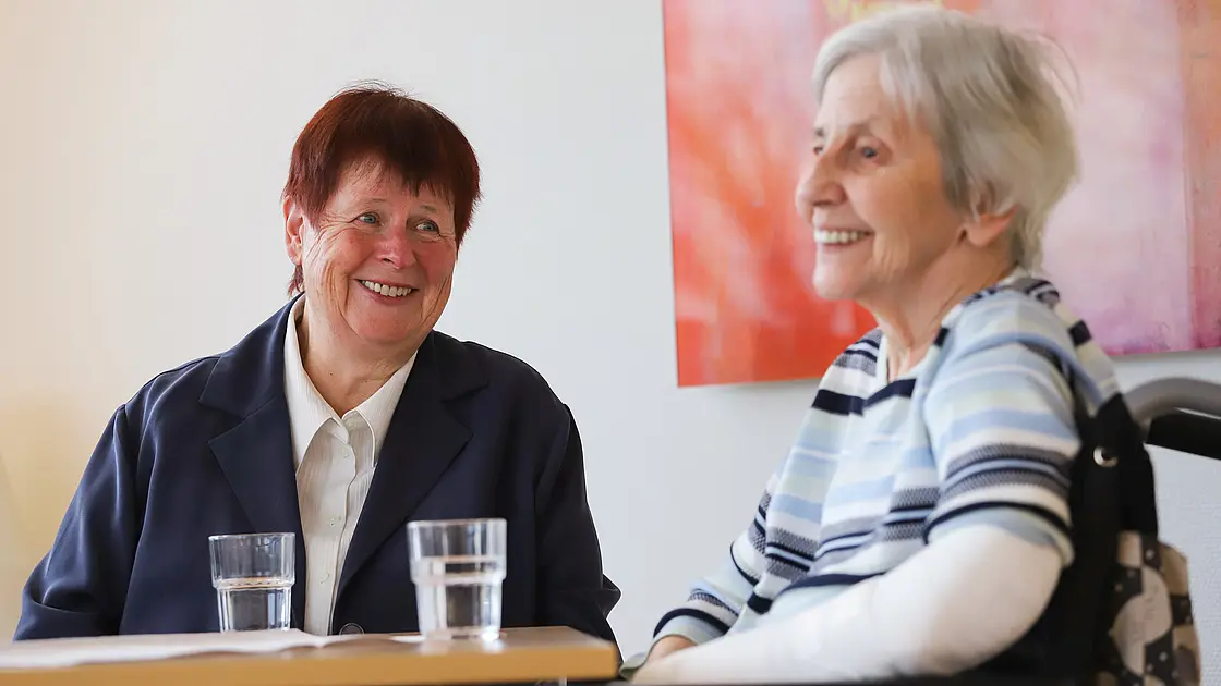 Zwei Personen sitzen an einem Tisch im Gespräch; die Szene wirkt ruhig und zugewandt.