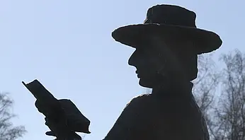 Dunkle Skulptur eines Mannes mit Hut, der ein Buch in den Händen hält, als Silhouette vor hellem Himmel dargestellt.