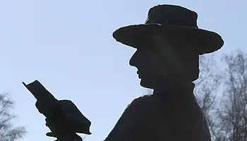 Dunkle Skulptur eines Mannes mit Hut, der ein Buch in den Händen hält, als Silhouette vor hellem Himmel dargestellt.