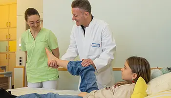 Dr. PD Daniel Windschall hält das angewinkelte Bein eines Kindes und untersucht den Fuß. Eine Mitarbeiterin steht daneben und beobachtet die Untersuchung.