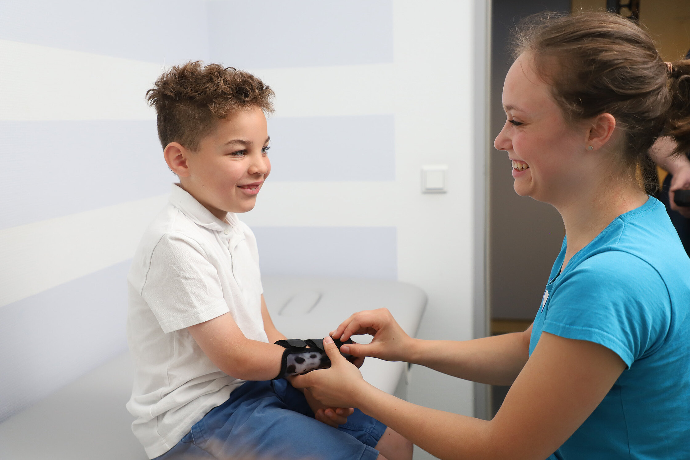 Ein Kind sitzt auf einer Untersuchungsliege, während eine Fachkraft eine Orthese am Unterarm anlegt und ausrichtet.