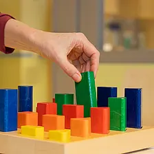 Eine Hand greift nach einem bunten Holzbaustein auf einem Steckbrett; verschiedene Formen und Farben sind geordnet angeordnet.