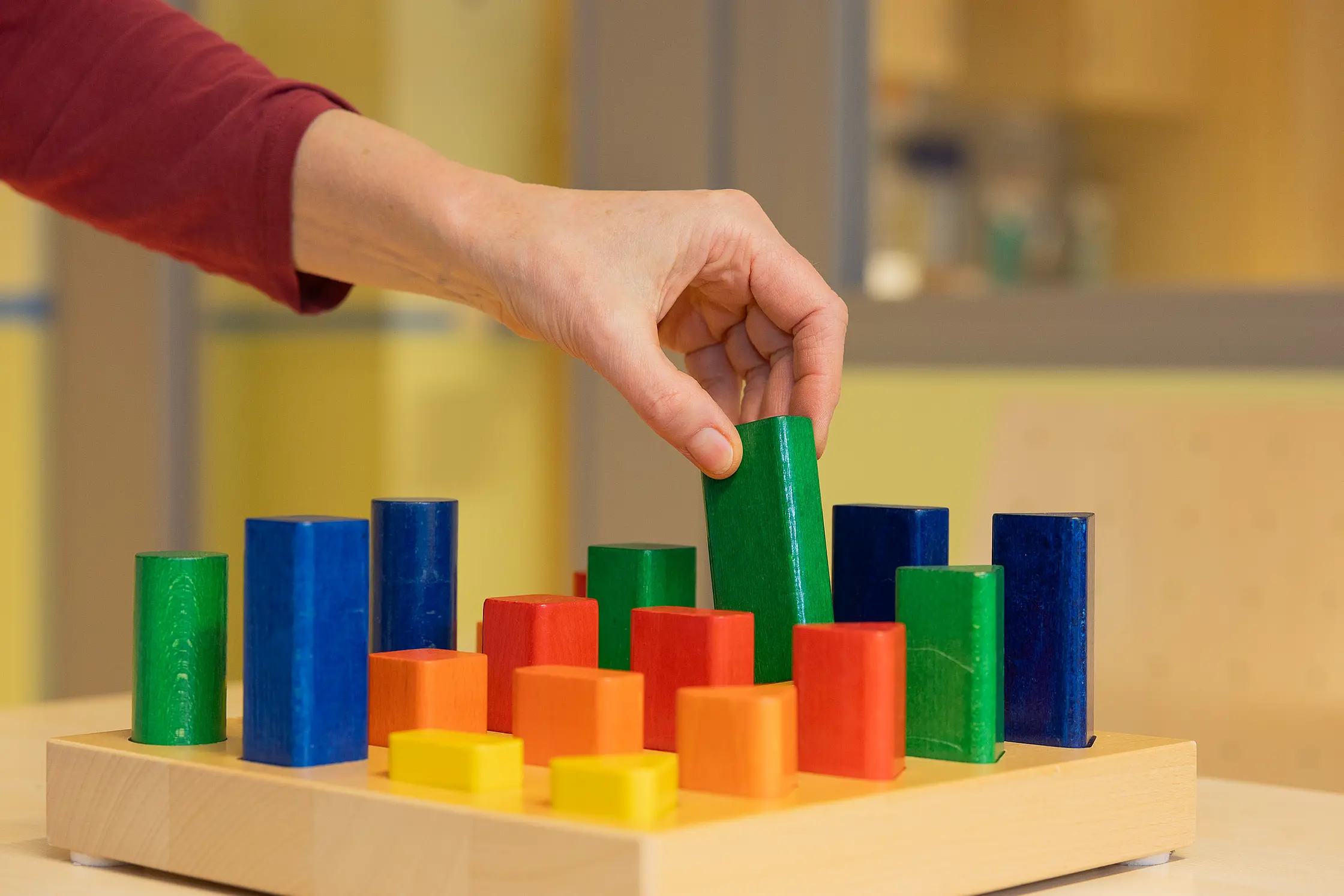 Eine Hand greift nach einem bunten Holzbaustein auf einem Steckbrett; verschiedene Formen und Farben sind geordnet angeordnet.