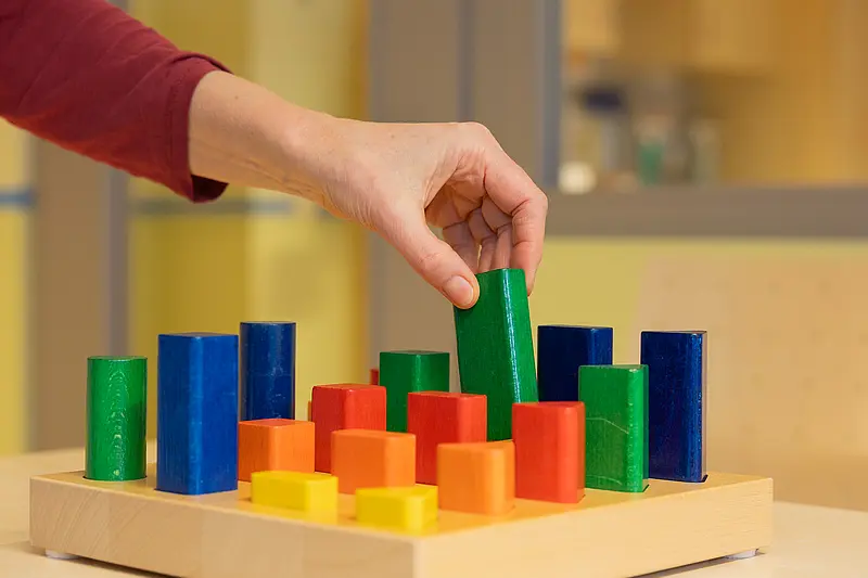 Mehrere bunte Holzformen stehen auf einem Steckbrett; die Anordnung dient dem Training von Feinmotorik und Koordination.