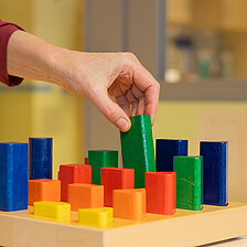 Eine Hand greift nach einem bunten Holzbaustein auf einem Steckbrett; verschiedene Formen und Farben sind geordnet angeordnet.