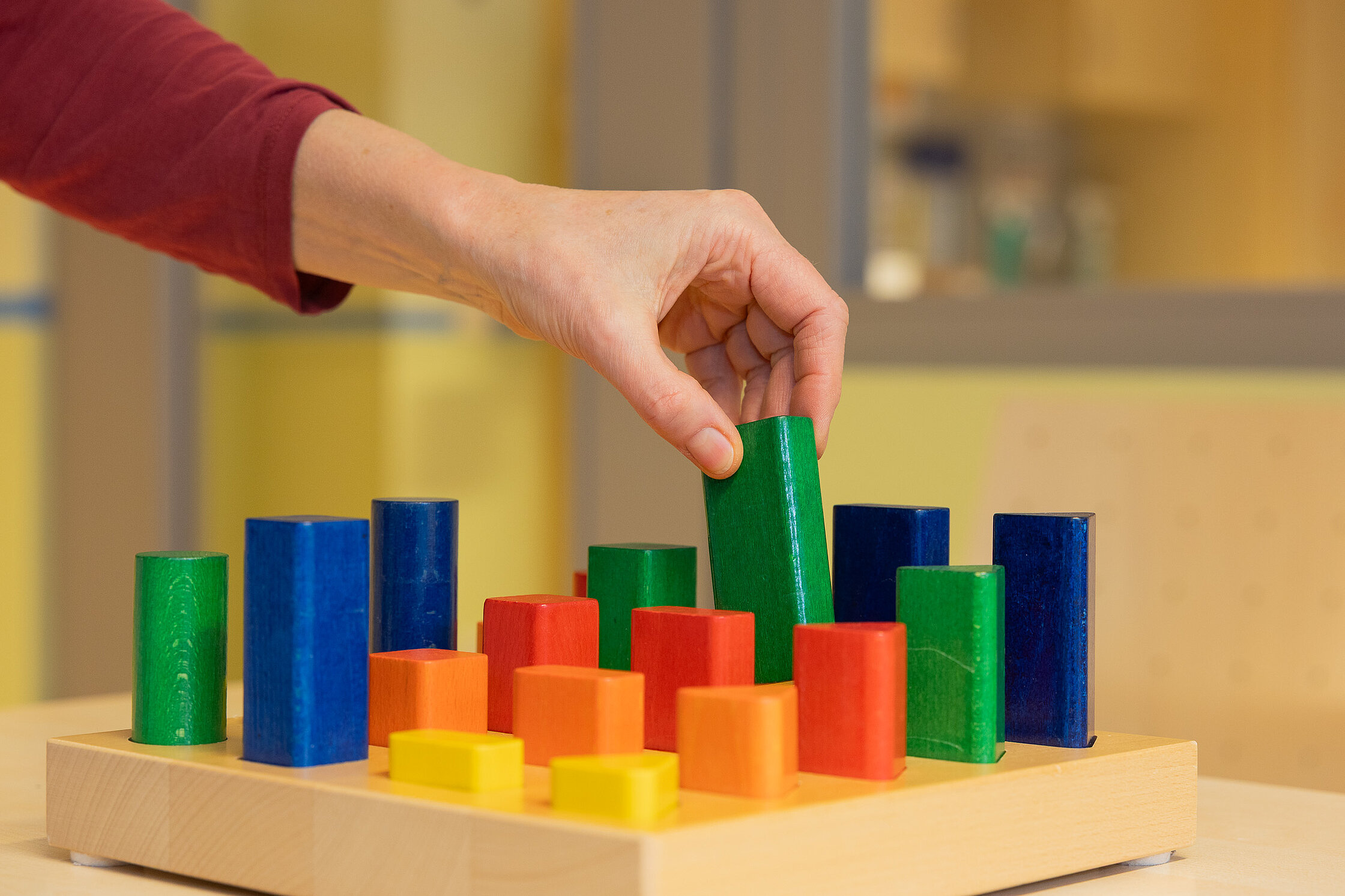 Eine Hand greift nach einem bunten Holzbaustein auf einem Steckbrett; verschiedene Formen und Farben sind geordnet angeordnet.