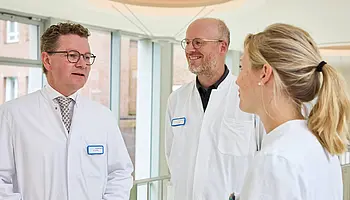 Dr. med. Ansgar Platte spricht im hellen Klinikflur mit zwei Mitarbeitenden in medizinischer Arbeitskleidung.