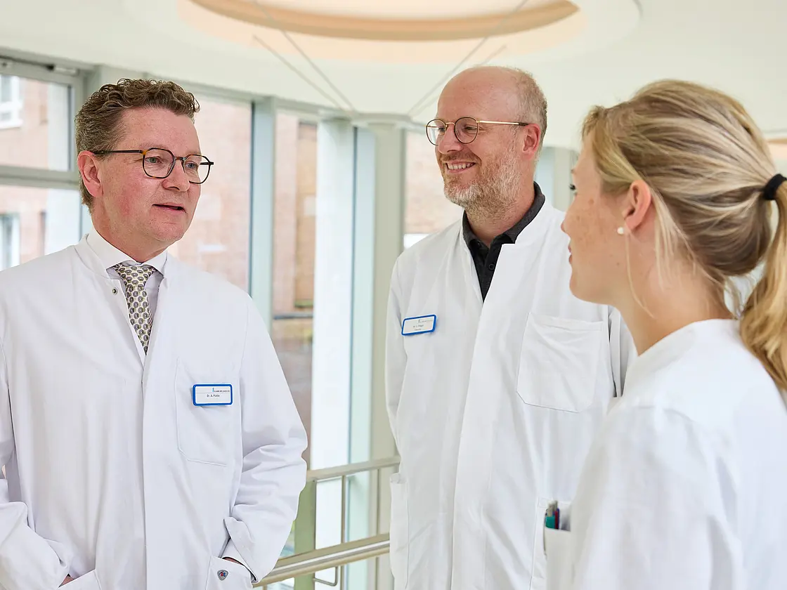 Dr. med. Ansgar Platte spricht im hellen Klinikflur mit zwei Mitarbeitenden in medizinischer Arbeitskleidung.