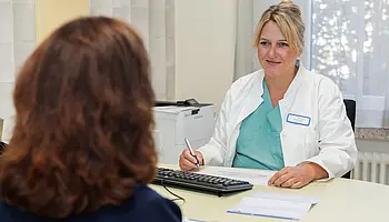 Ärztin sitzt an einem Schreibtisch und spricht mit einer Patientin; vor ihnen liegen Unterlagen und ein Computer.