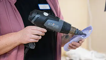 Nahaufnahme einer ergotherapeutischen Arbeitssituation: Eine Fachkraft erwärmt mit einem Heißluftgerät thermoplastisches Material, um eine Handschiene individuell zu formen; das Material wird in der Hand gehalten und bearbeitet.