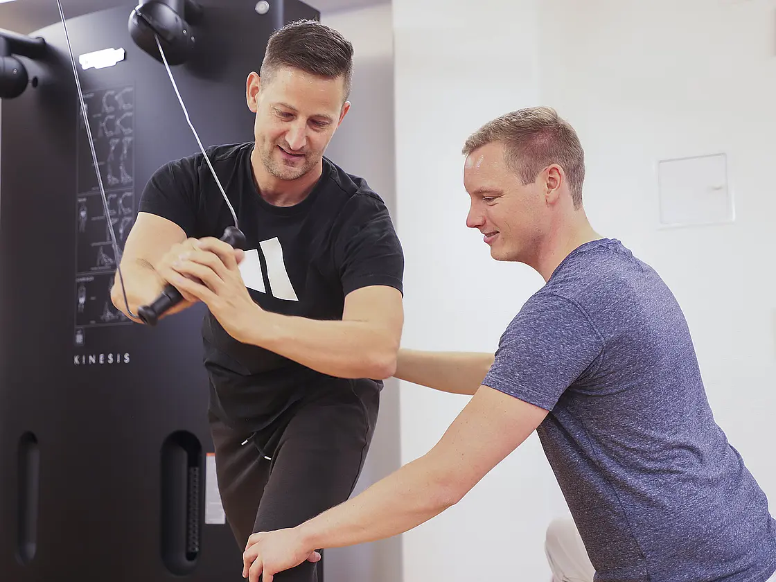 Ein Patient führt eine Übung an einem Trainingsgerät aus, während ein Therapeut unterstützend an seiner Seite steht.