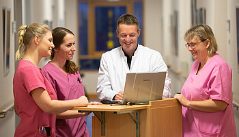 Ärztlicher Mitarbeiter und Pflegekräfte stehen im Flur an einem Stehpult und besprechen Informationen am Laptop.